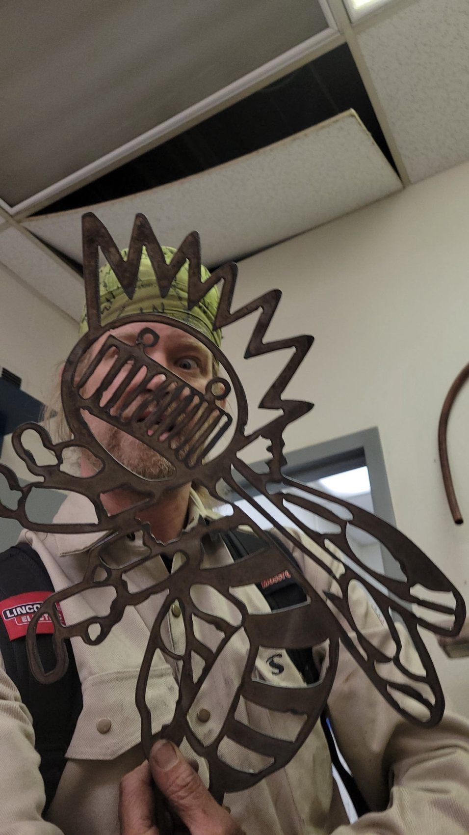 The maker in a Lincoln Electric welding jacket holding up a hand-cut steel bee emblem in the shop